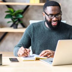 Cheerful African-American male student or worker in glasses is watching online lectures or webinars and writing notes in a notebook
