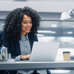 Modern Office: Black Businesswoman Sitting at Her Desk Working on a Laptop Computer. Smiling Successful African American Woman working with Big Data e-Commerce. Motion Blur Background